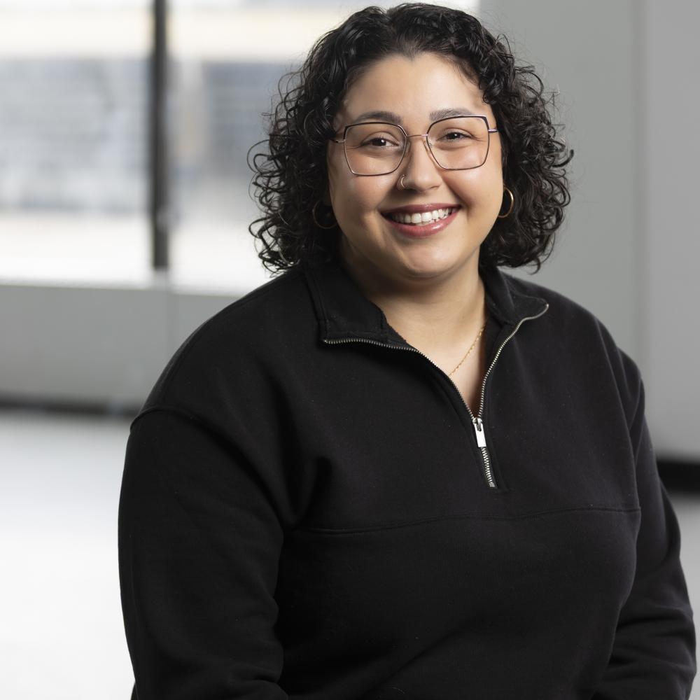 A woman with short curly hair is seen smiling toward the camera with a black quarter-zip sweatshirt. A window is seen blurred in the background.