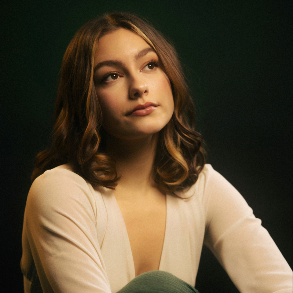 A white woman lifts her face towards a soft light. She has lightly curled long brown hair and wears a white long sleeve shirt.