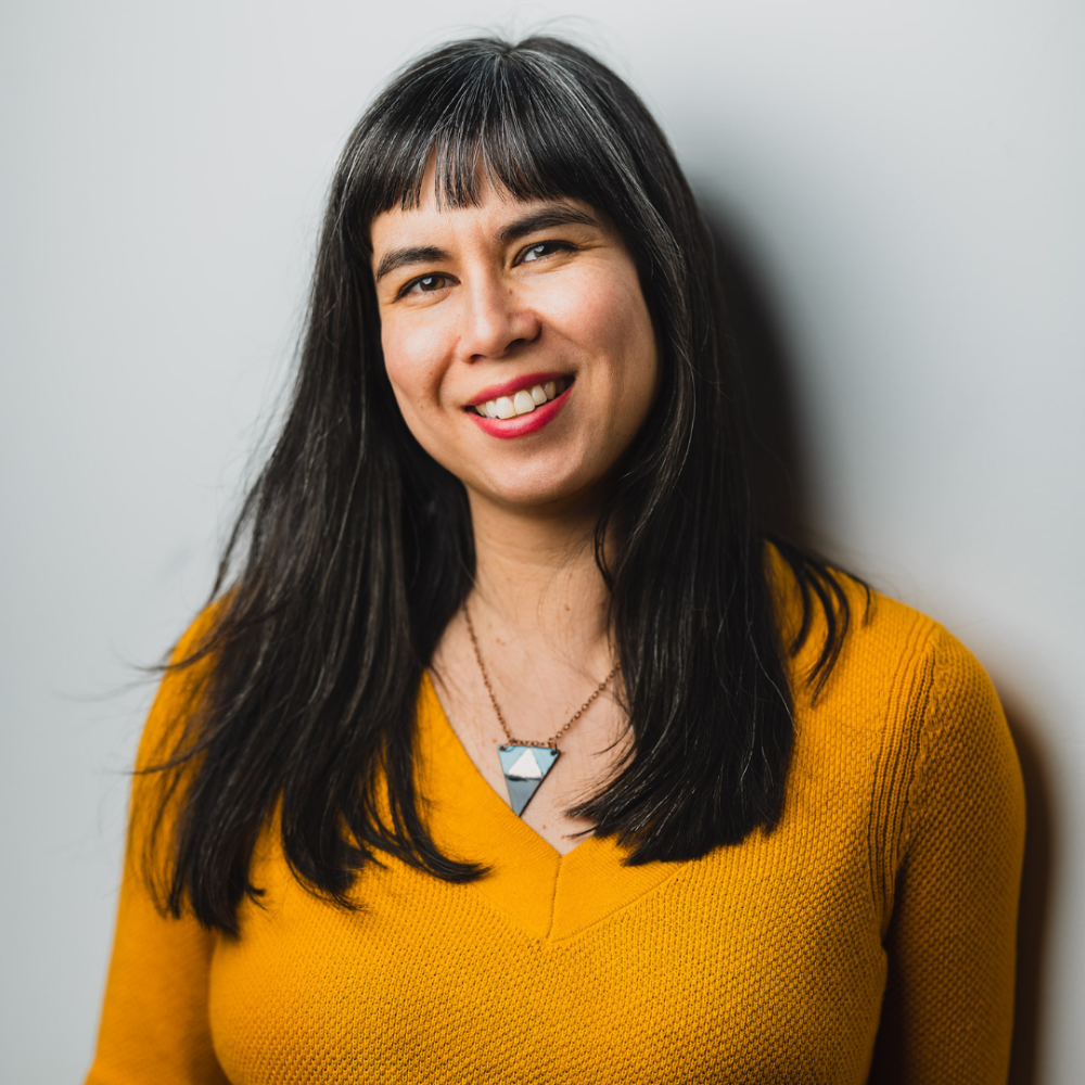A Chinese-Canadian woman stands against a white wall smiling. She has long black hair with bangs. She is wearing a mustard long sleeve short and has a triangle necklace.