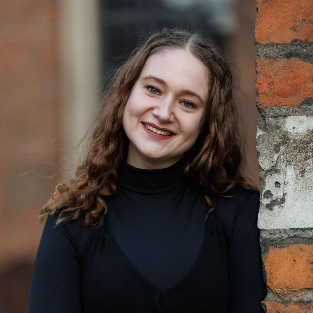 A white woman with blue eyes and long curly hair is seen leaning on a brick wall from the chest up. She is wearing a black turtleneck, and the background is blurred.
