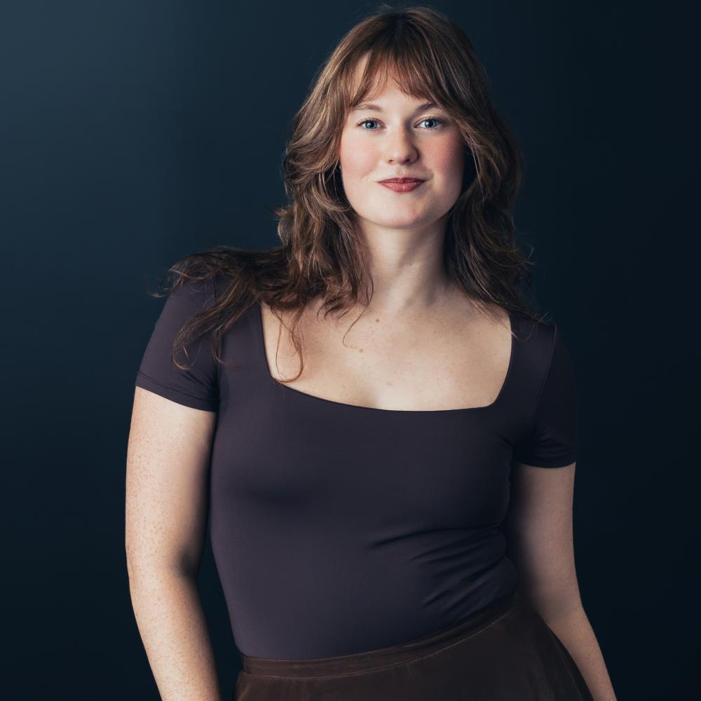 A white woman with long curly Auburn hair is seen against a dark background. She is wearing a dark purple square cut t-shirt and is smiling toward the camera.