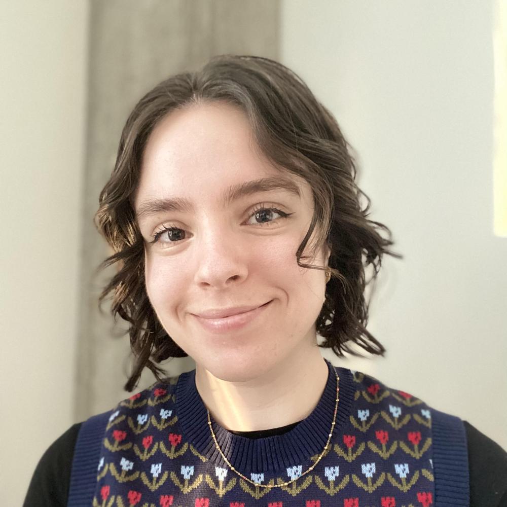 A woman with light skin and short curly hair is smiling toward the camera. She is wearing a flower printed sweater and a gold necklace. The background of the photo is beige and grey.