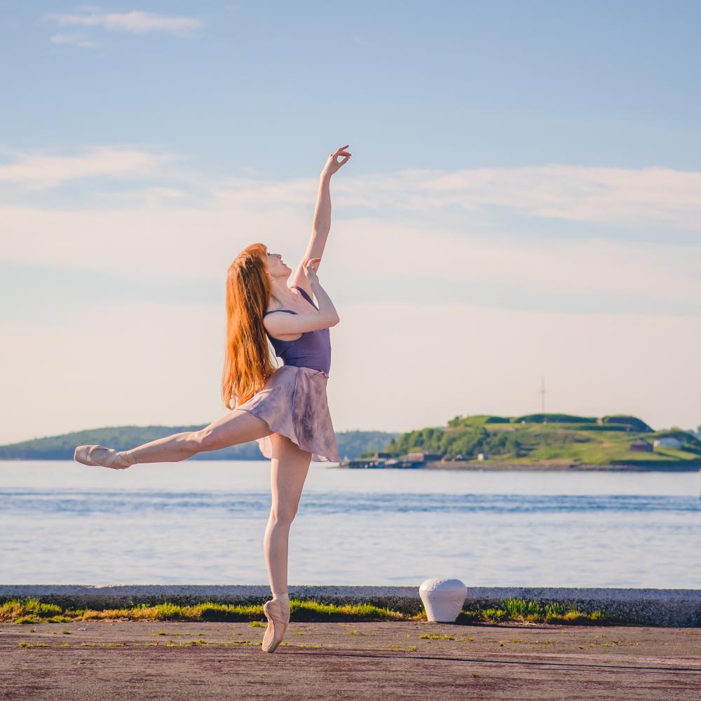 A woman with light skin and red hair is seen wearing a purple leotard and light wrap skirt. She is balanced on one leg, with the other extended wearing pointe shoes. Her gaze looks upwards to her extended arm. In the background blue sky and ocean are seen and an island.