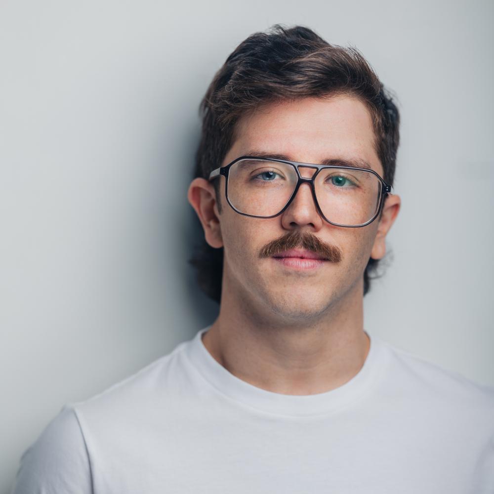 A white man with a brown mustache, mullet and glasses is seen wearing a white shirt against a grey background. They are looking towards the camera with a soft expression.
