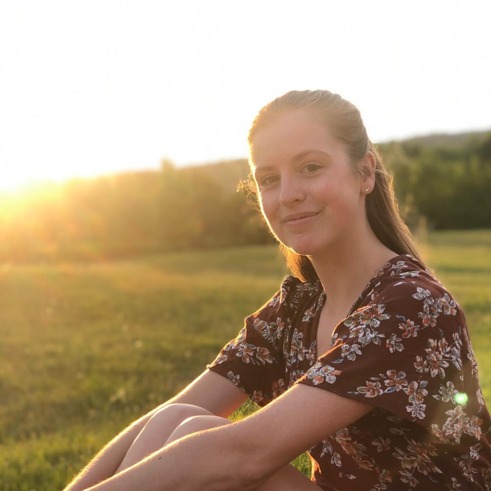 A white woman with long light brown hair is seen sitting in a field smiling toward the camera. She is wearing a burgundy top with white flowers. Trees and the setting sun are seen in the background. 