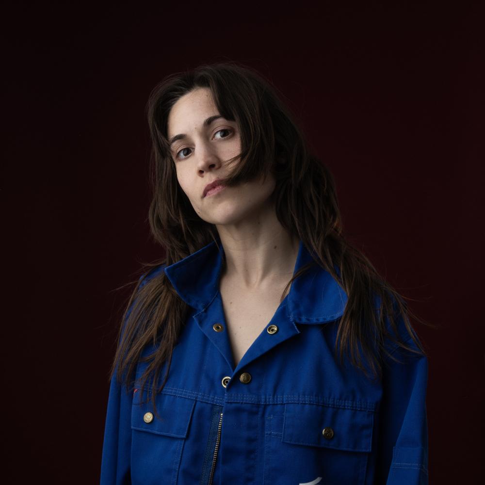 A dancer with light skin and long dark hair wears a blue jacket against a black background. Their head is tilted to the left. 